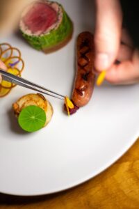 Gros plan d'un chef dressant avec précision un plat gastronomique composé de saucisses et de légumes.