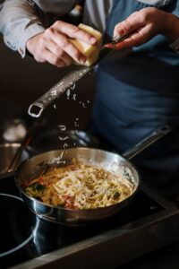 Un chef en tablier râpe du fromage sur des pâtes dans une cuisine moderne, capturant l'art culinaire en action.